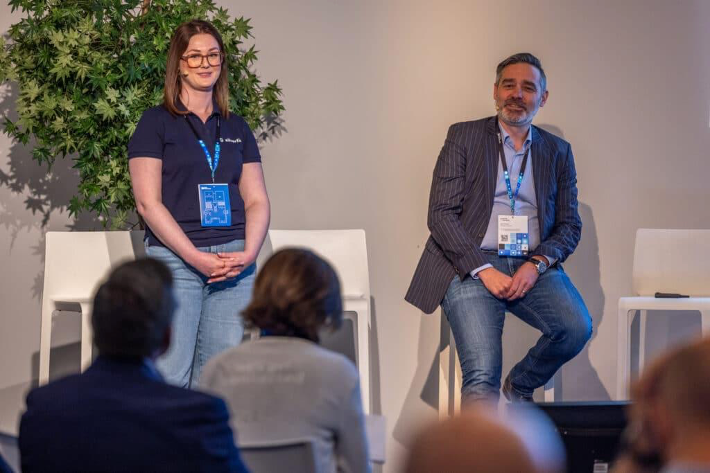 Two individuals seated on chairs at a conference, engaged in a discussion amidst a professional setting.